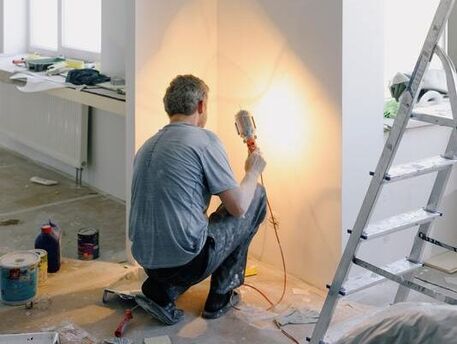 Man in gray shirt kneeling beside wall with light to inspect construction work in room being remodeled